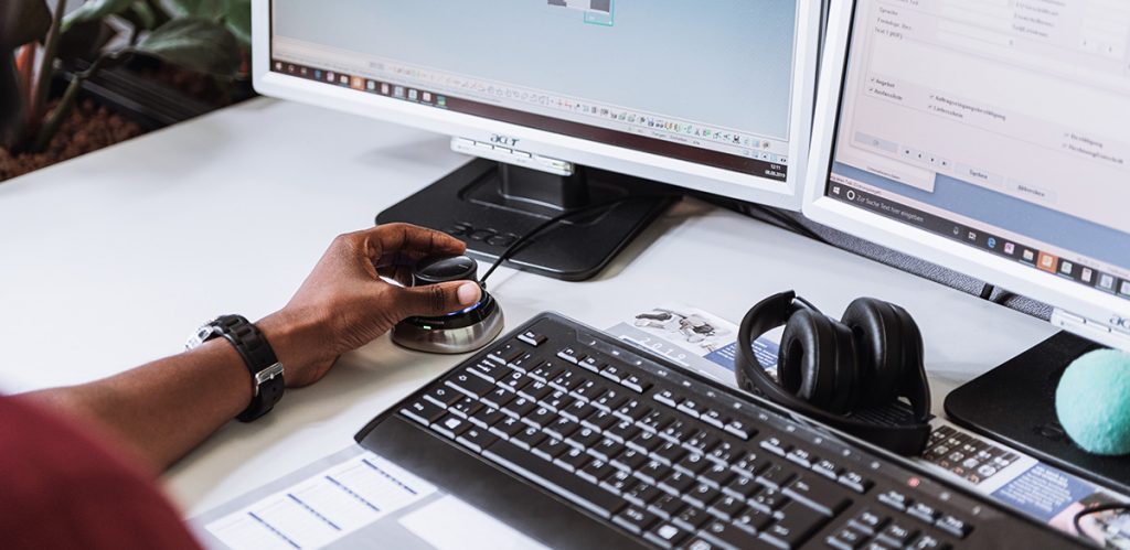 Schreibtisch mit Tastatur und zwei Bildschirmen. Die linke Hand einer Person bedient eine Maus. Vor der Tastatur liegen Kopfhörer.